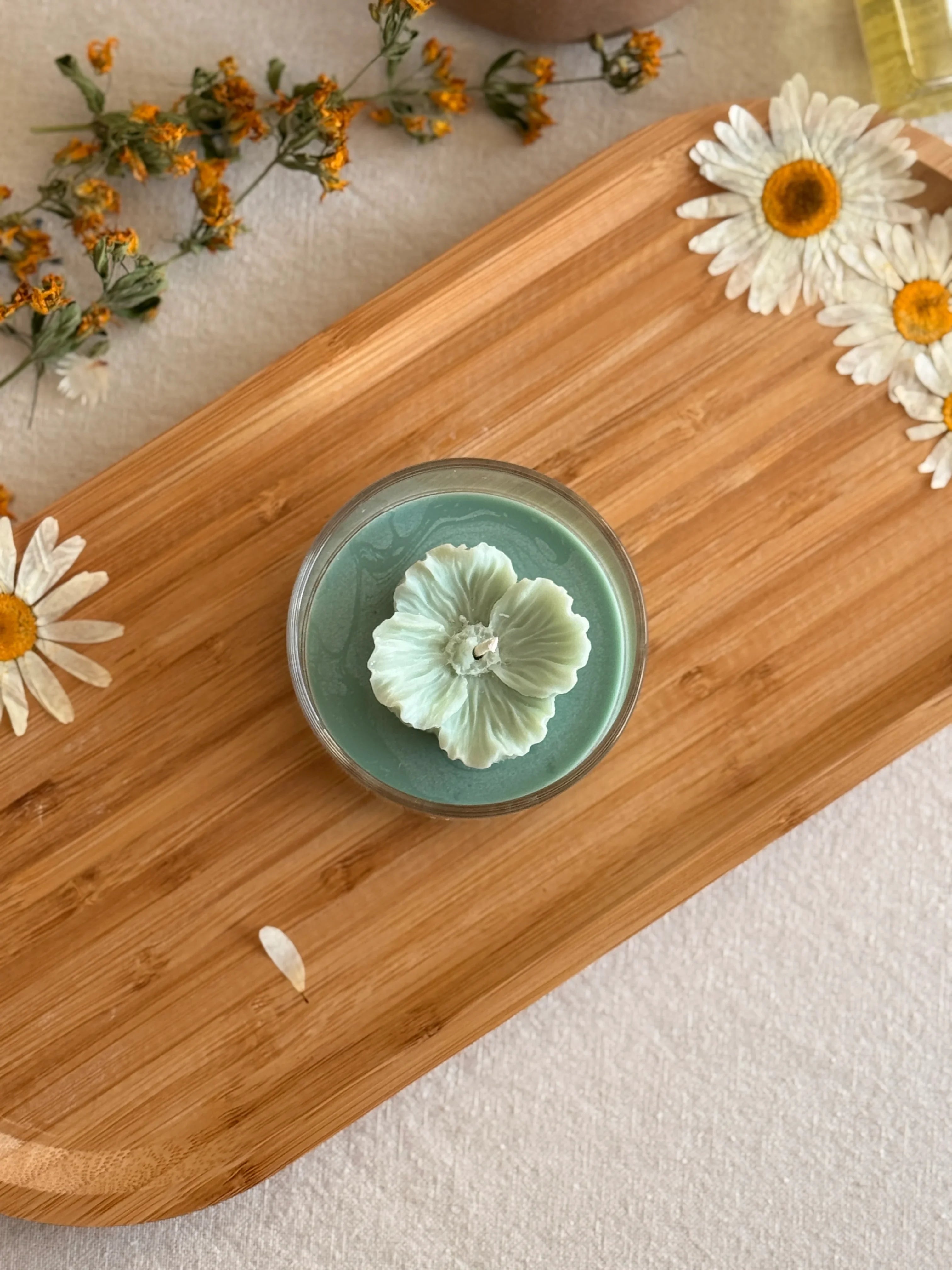 Handmade soy candle with flower design on wooden tray, surrounded by dried daisies and herbs for cozy home decor