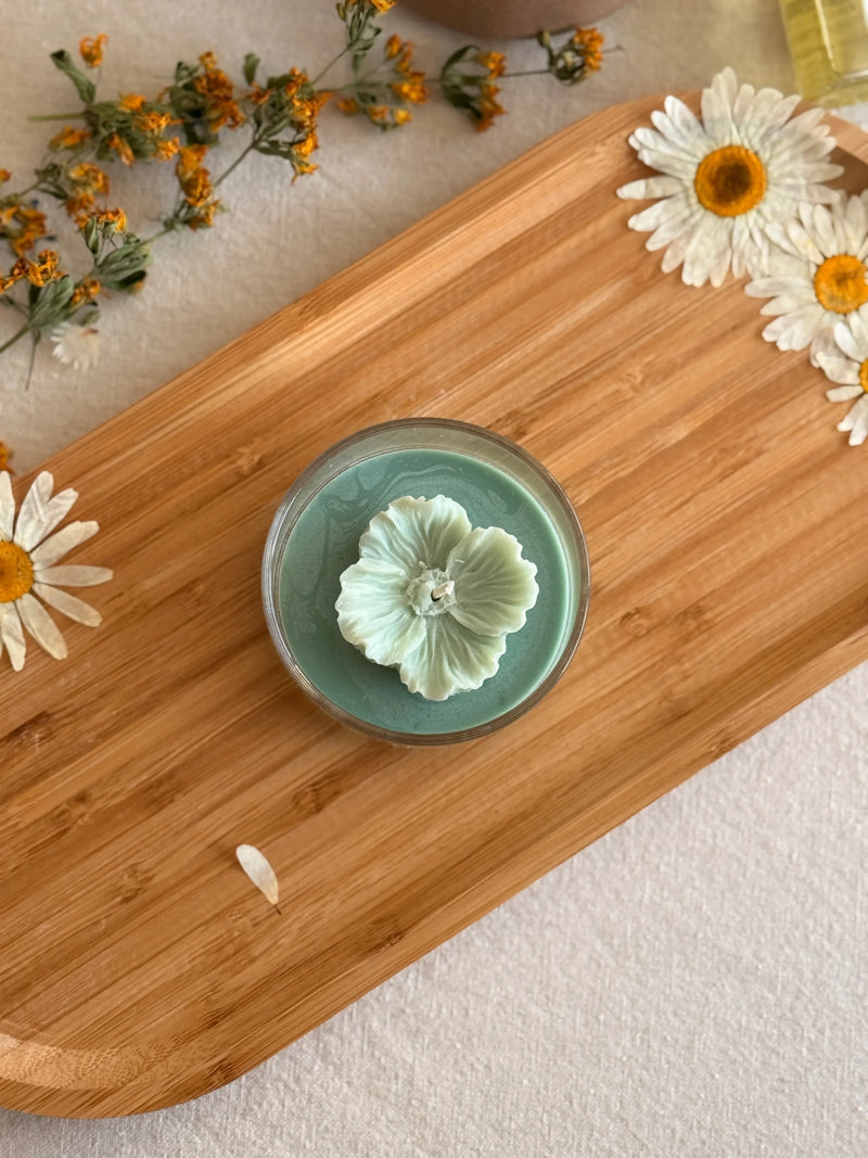 Handmade soy candle with flower design on wooden tray, surrounded by dried daisies and herbs for cozy home decor