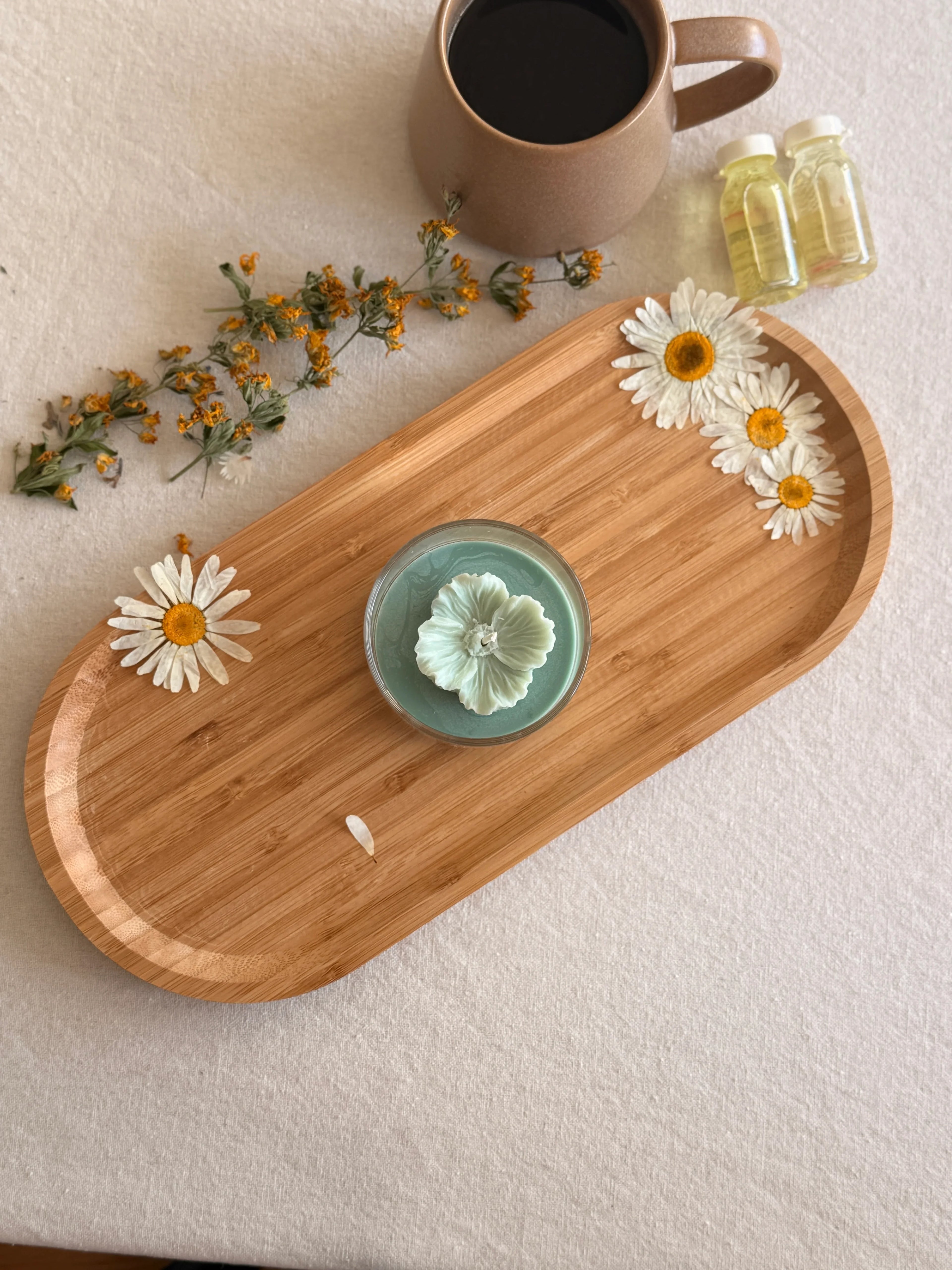 Handmade soy candle with flower design on wooden tray, surrounded by dried chamomile, coffee mug, and essential oils for cozy home décor.
