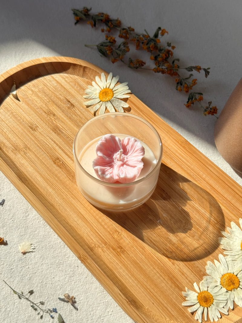 Handmade soy candle with pink flower design on wooden tray, surrounded by dried daisies for cozy home decor.