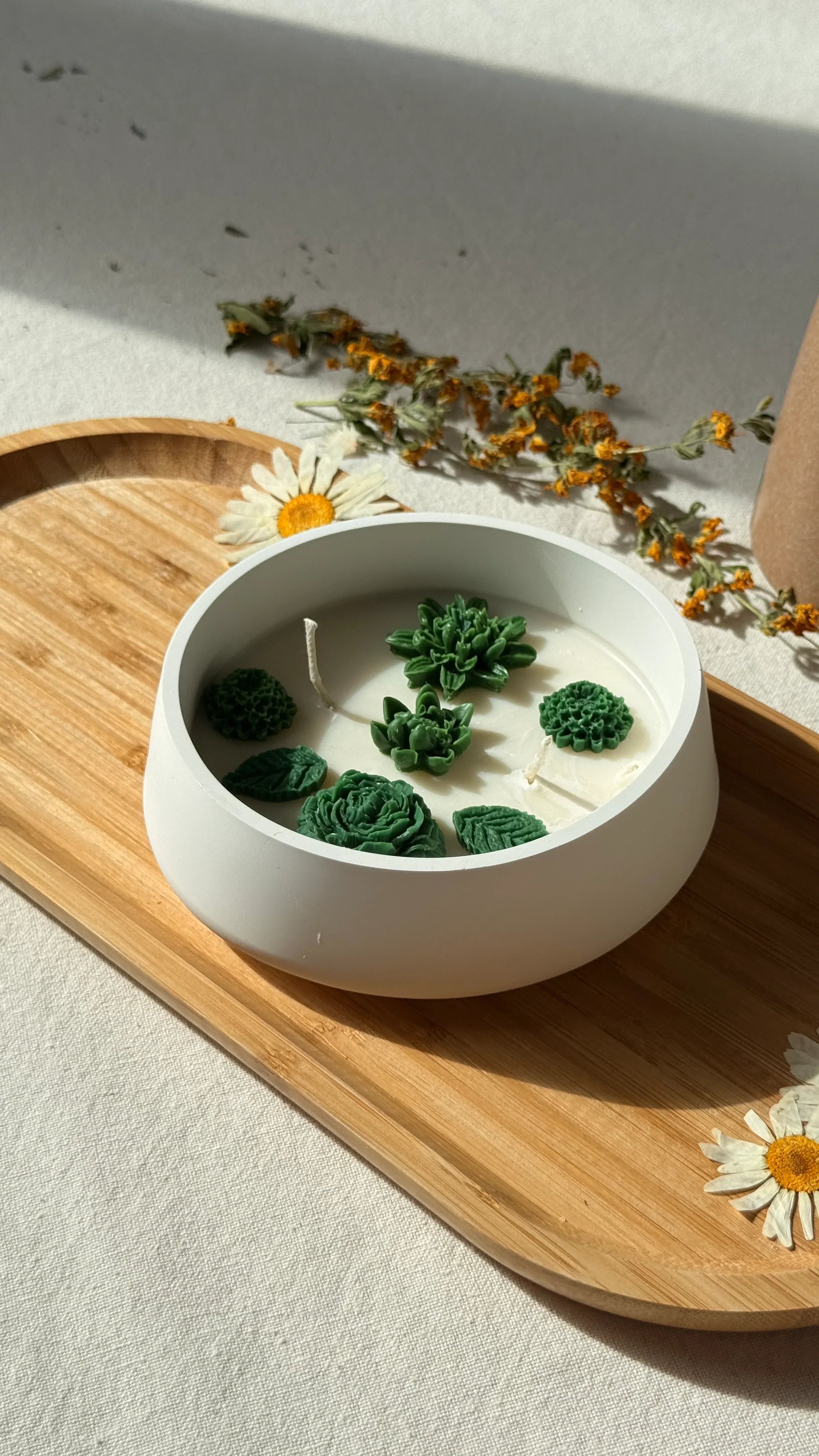 Handmade soy candle with green succulent wax decor in white bowl, styled on wooden tray with dried flowers for cozy home ambiance.