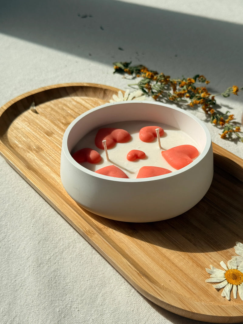 Handmade soy candle with coral hearts in white ceramic bowl, styled on wooden tray with dried flowers for cozy home décor.