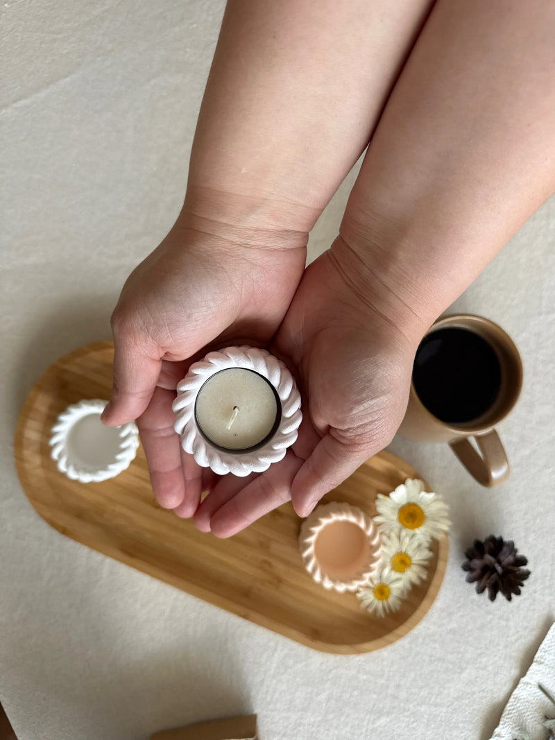 Hands holding a white scalloped soy candle with wooden tray, daisies, and coffee mug for cozy Scandinavian home décor