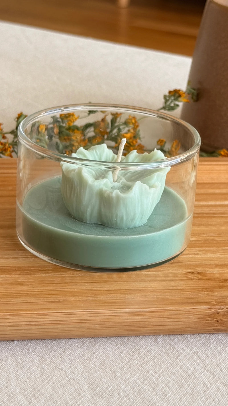 Handmade soy candle in clear glass jar with ruffled green wax, set on wooden surface with dried flowers.