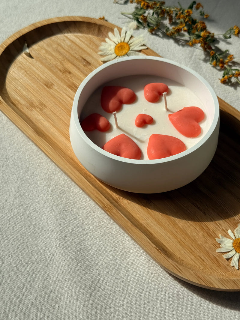 Handmade soy candle with coral hearts in white ceramic bowl, styled on wooden tray with dried flowers for cozy home décor.