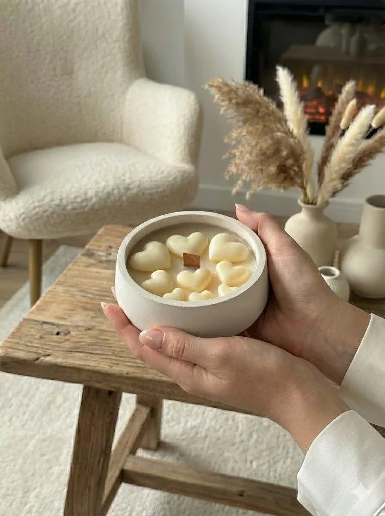 Hands holding a ceramic bowl of heart-shaped soy wax melts, cozy Scandinavian home décor with pampas grass and fireplace