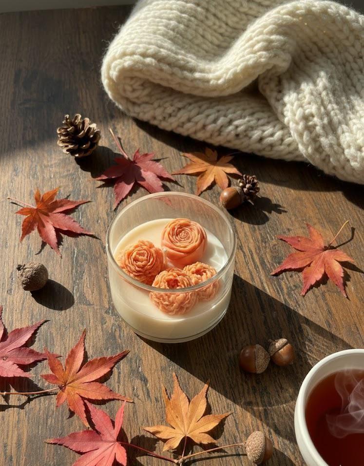 Handmade soy candle with rose-shaped wax, autumn leaves, pinecones, and cozy knit blanket on wood